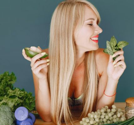 A smiling woman holding avocado and basil, surrounded by fresh vegetables and fruits for healthy eating.