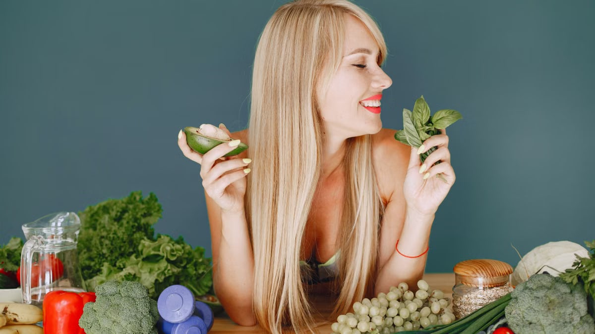 A smiling woman holding avocado and basil, surrounded by fresh vegetables and fruits for healthy eating.
