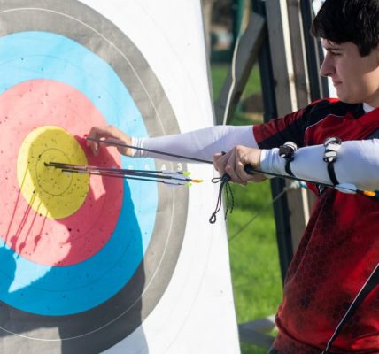Focus on man aiming with bow and arrows at target during outdoor archery practice.