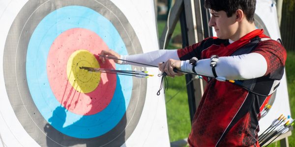 Focus on man aiming with bow and arrows at target during outdoor archery practice.