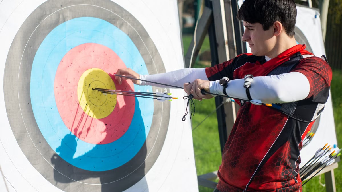 Focus on man aiming with bow and arrows at target during outdoor archery practice.